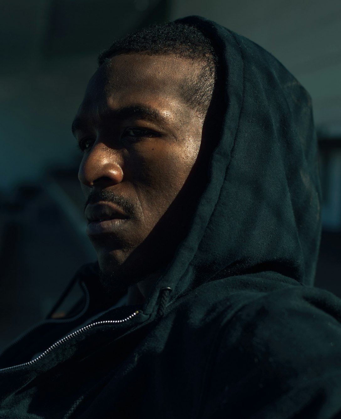 Close-up portrait of an African American adult male with a hooded jacket, captured indoors with dark lighting creating a dramatic mood.