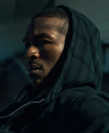 Close-up profile of a black man wearing a dark hooded sweatshirt under moody directional lighting by Brian Lowe.