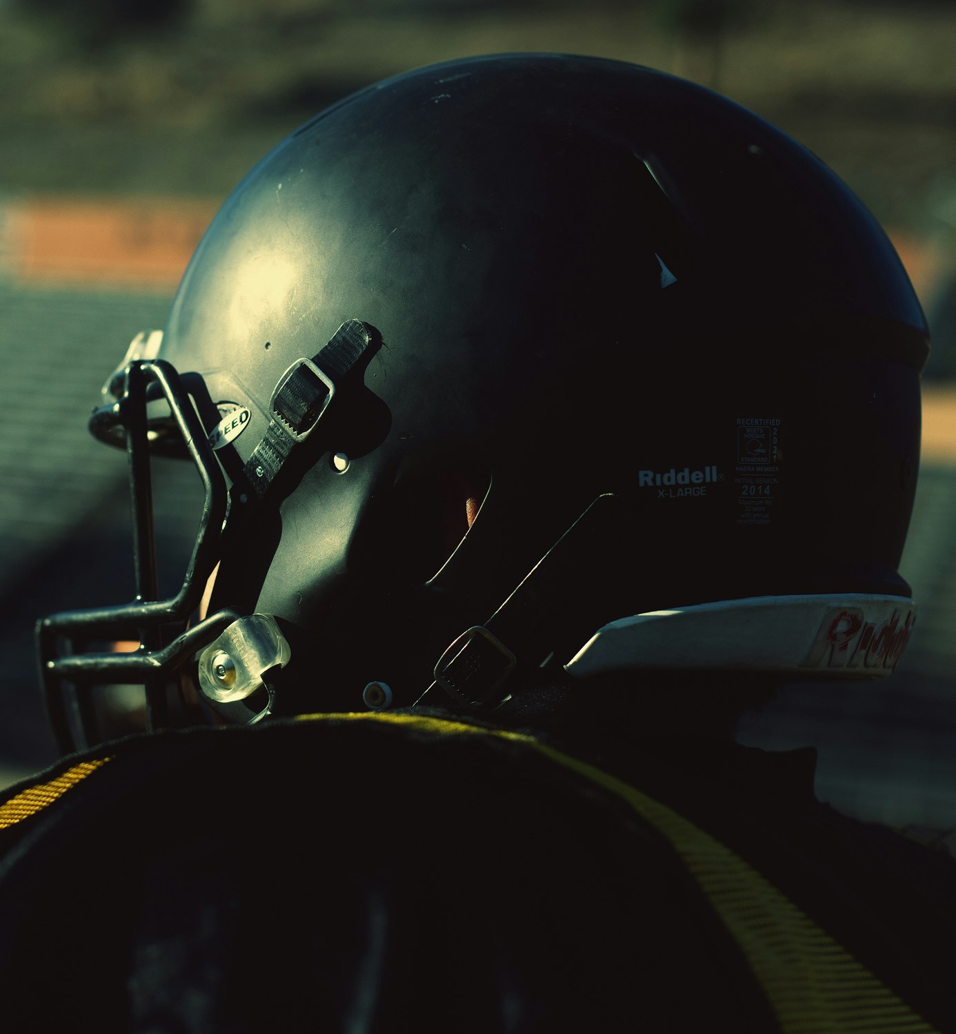 Close-up of a football helmet in black with a face mask, showing the side with manufacturer's label against an indistinct background.