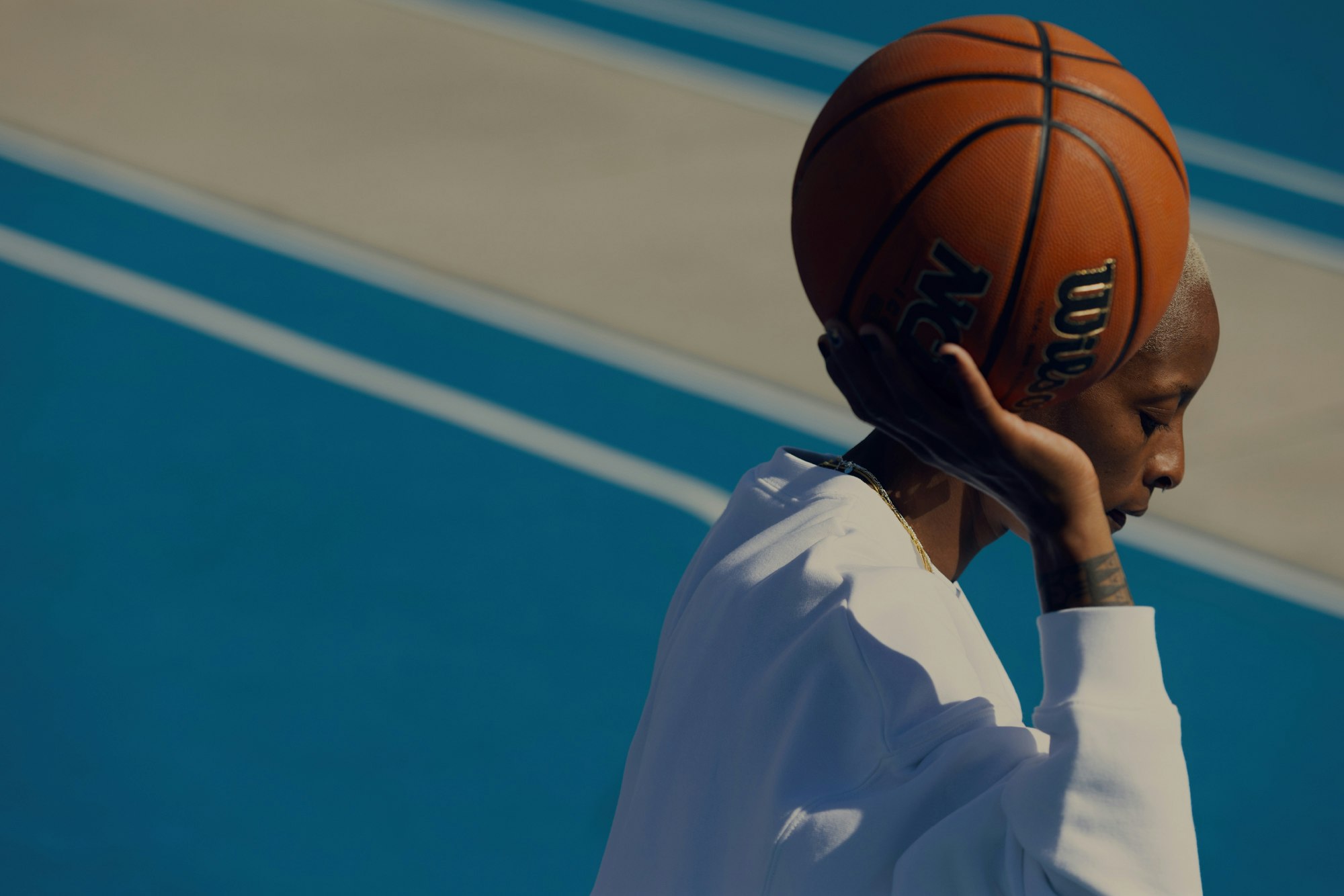 A young adult with short hair rests her head against a basketball on a blue sports court, wearing a white sweatshirt and gold necklace.
