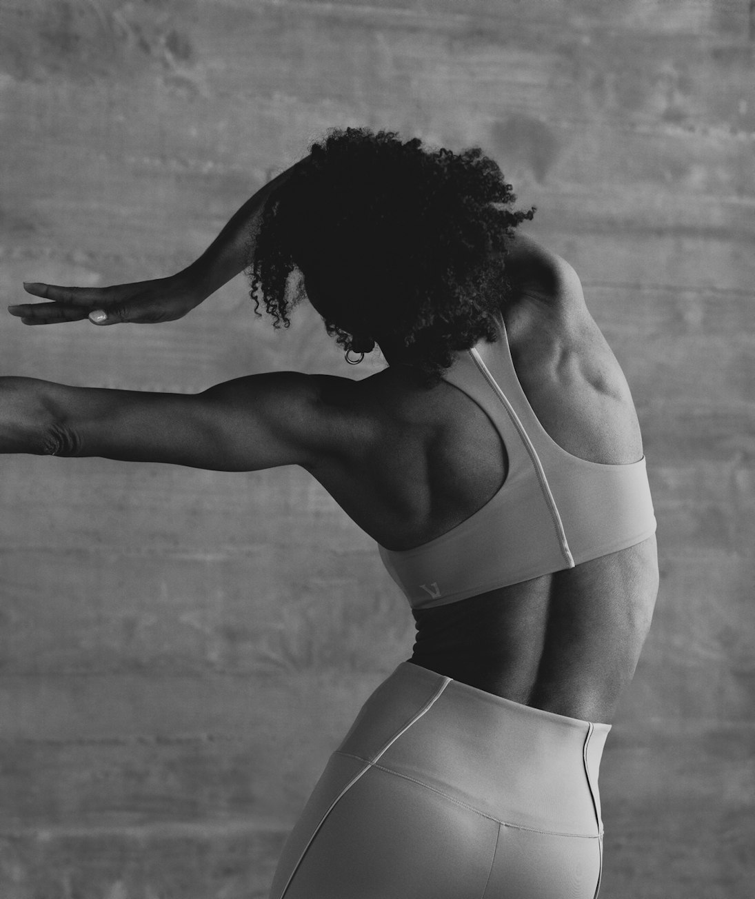 Athletic African-American woman in workout gear poses in front of a grey textured wall, showcasing fitness and flexibility in a studio setting.