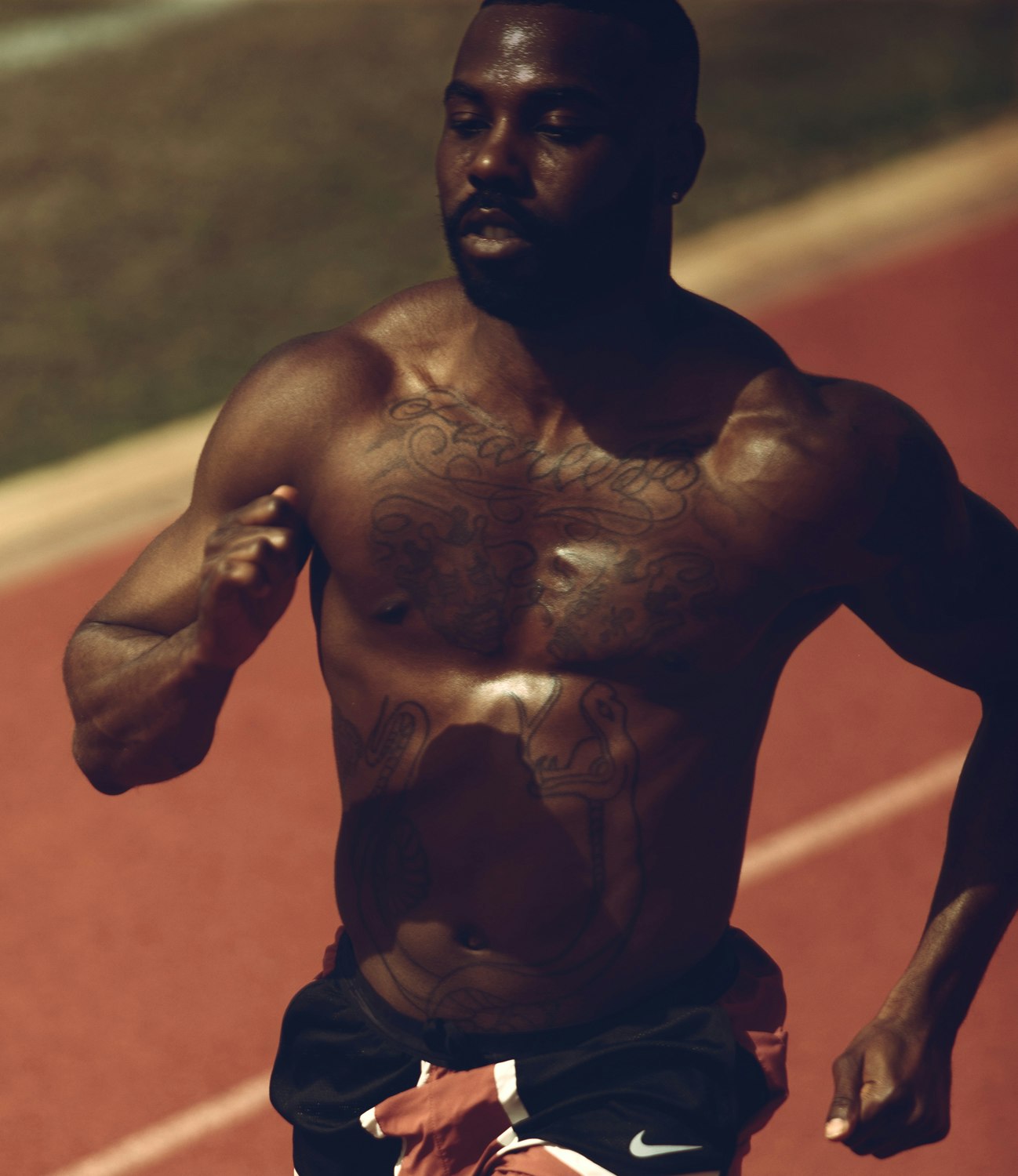 Muscular shirtless man running on an outdoor track on a sunny day, his brow furrowed in determination, wearing athletic shorts and Nike shoes.