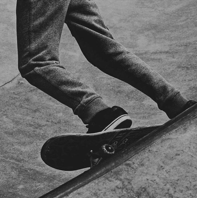 A close-up, black and white shot of a skateboarder's legs and feet on a skateboard, angled towards a rail in a skate park.