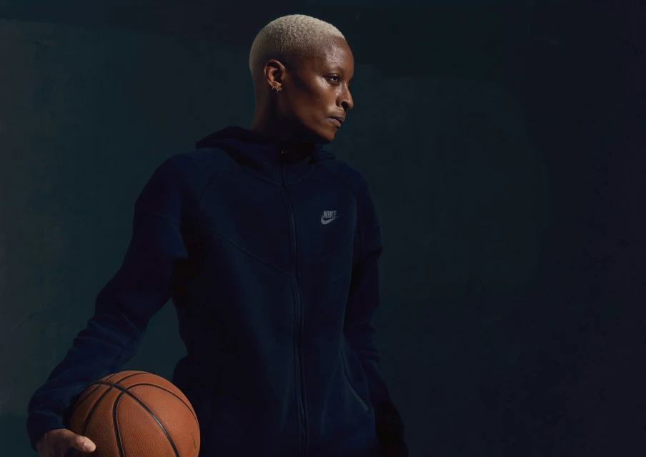Adult female athlete in sportswear holding a basketball against a dark blue backdrop, looking to the side in a fitness studio.