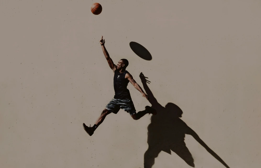 African American man jumps to make a basketball shot against a beige wall, his long shadow visible, athletic, wearing a tank top.