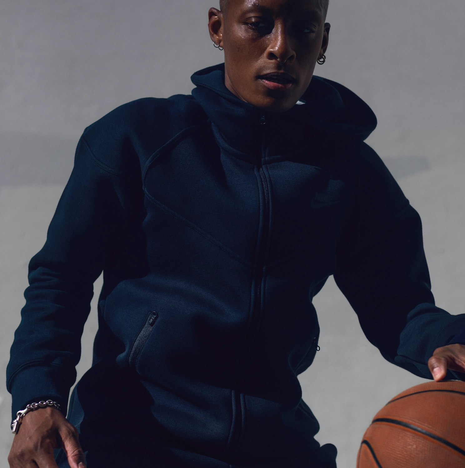 Adult male athlete in a dark-blue hooded sweatshirt dribbling an orange basketball in a studio with a neutral-gray background.