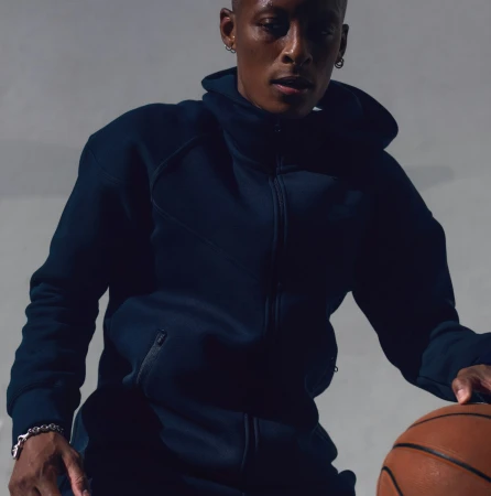Adult male athlete in a dark-blue hooded sweatshirt dribbling an orange basketball in a studio with a neutral-gray background.