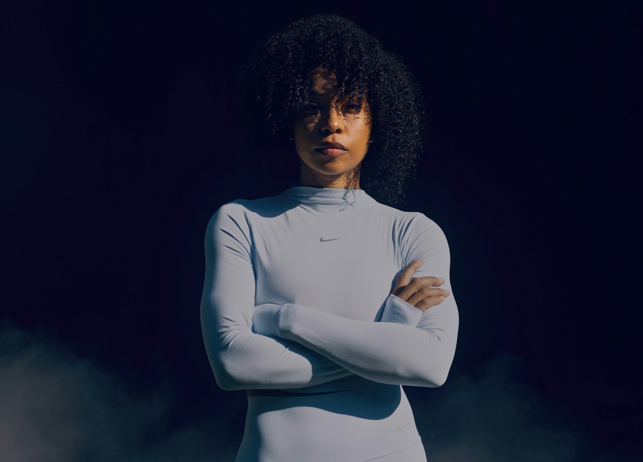 Portrait of a woman with dark curly hair wearing a long-sleeve white top with arms crossed against a dark background.