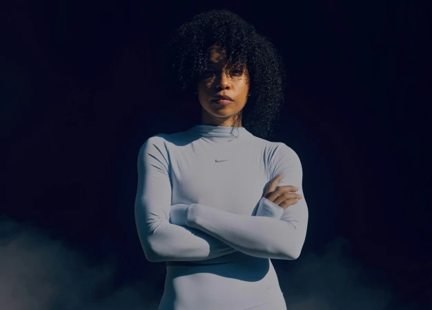 Portrait of a woman with dark curly hair wearing a long-sleeve white top with arms crossed against a dark background.