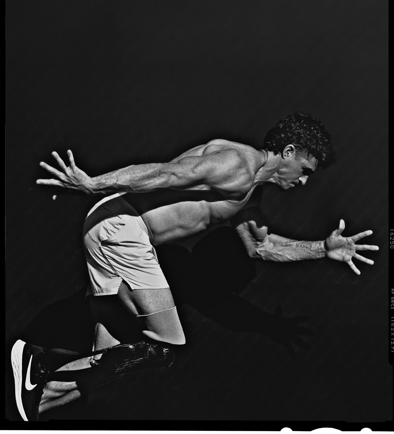 Monochrome shot of a muscular man with a prosthetic leg in running pose, wearing shorts and Nike shoes against a dark background.
