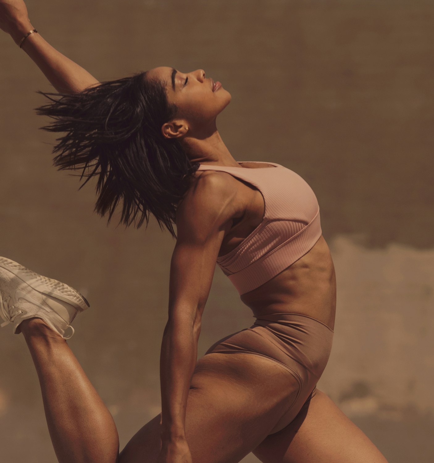 Athletic woman in sportswear leaping with outstretched arm against a neutral background, showcasing fitness and active lifestyle in motion.