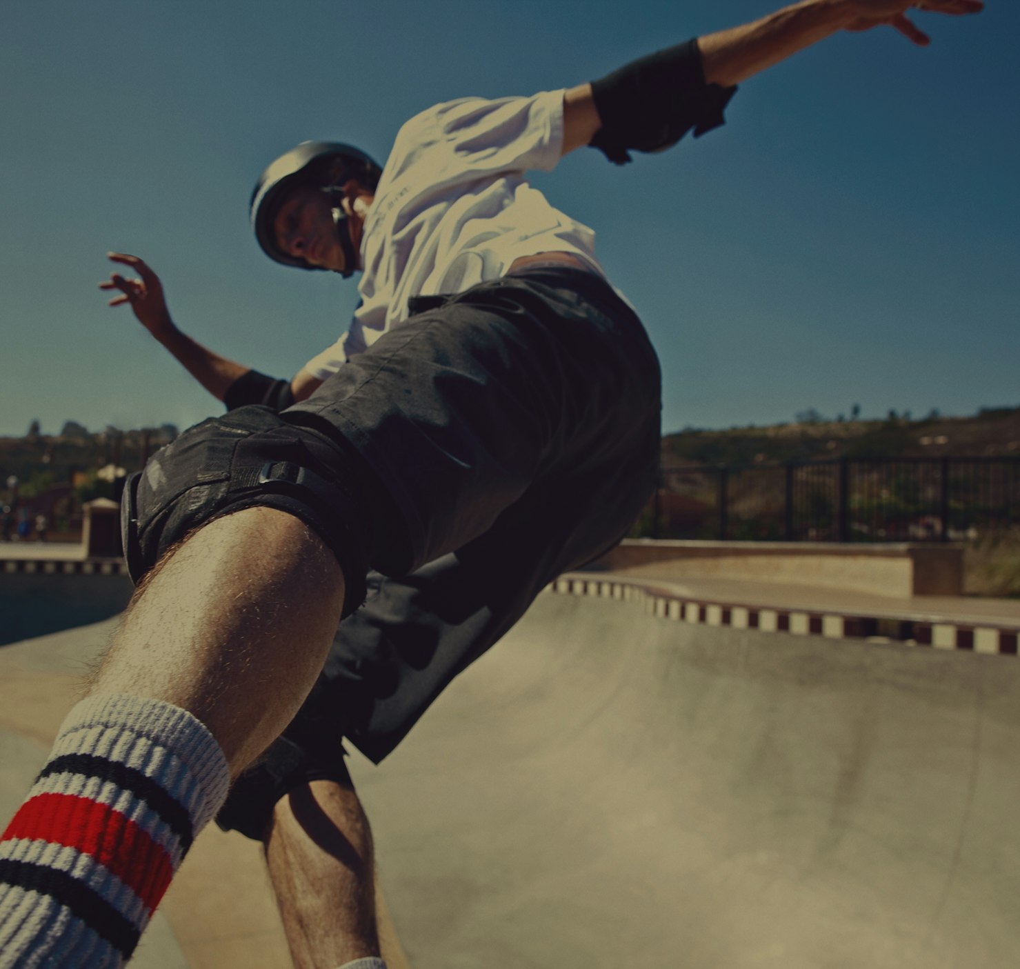 A skater in mid-air wearing a helmet and pads. A skatepark bowl, blue sky, and distant trees fill the background on a sunny day.
