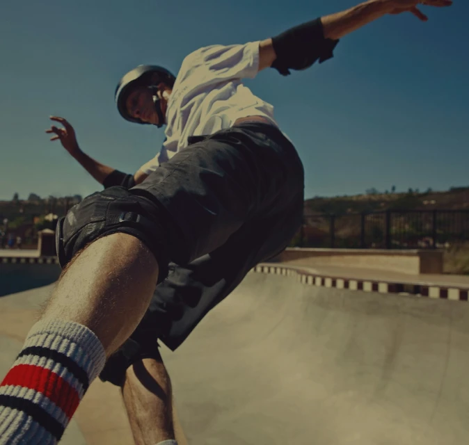A skater in mid-air wearing a helmet and pads. A skatepark bowl, blue sky, and distant trees fill the background on a sunny day.