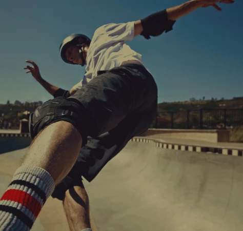 A skater in mid-air wearing a helmet and pads. A skatepark bowl, blue sky, and distant trees fill the background on a sunny day.