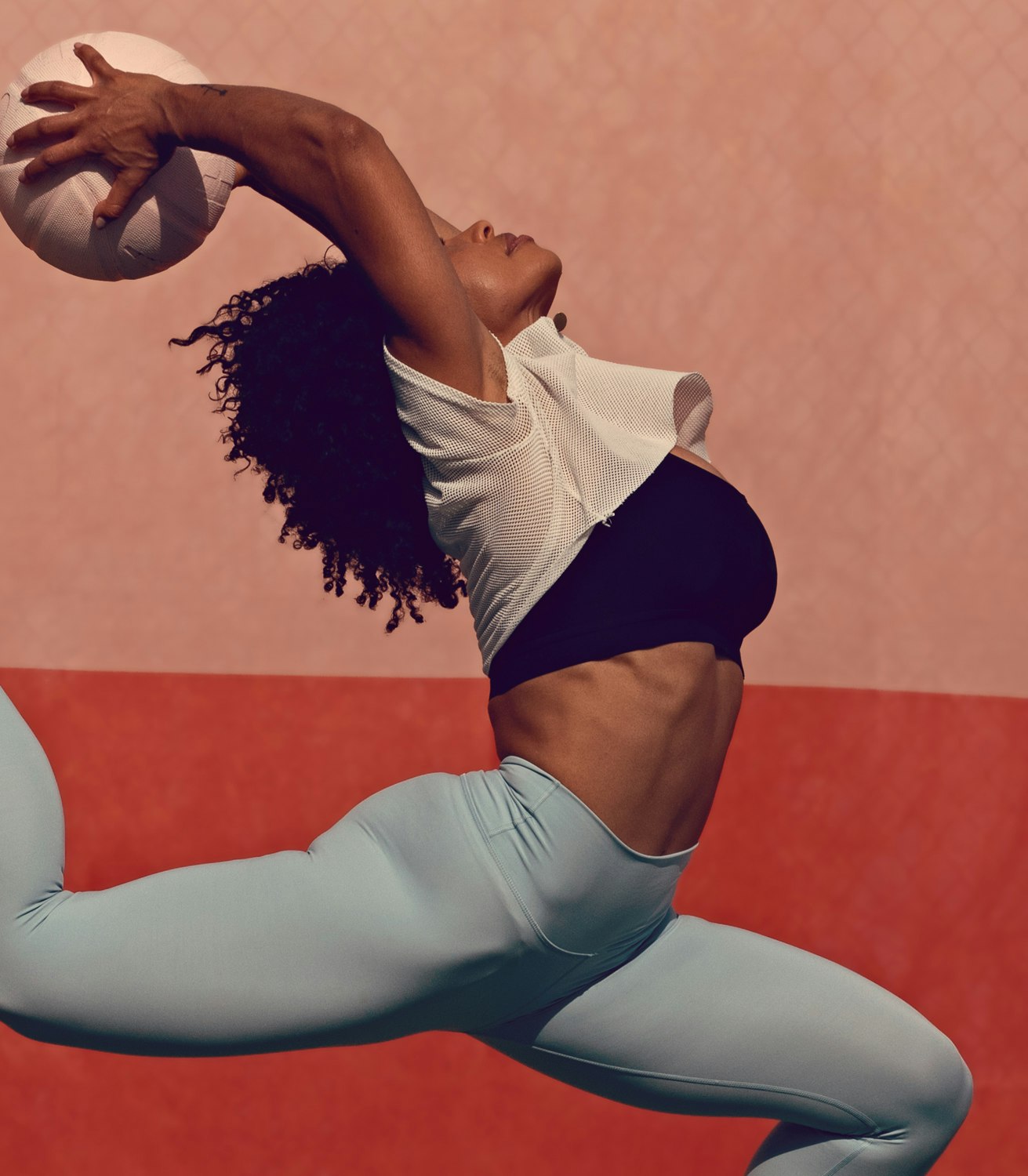 A woman in athletic wear leaps against a split pink and orange background, reaching to catch a white ball with one hand overhead.