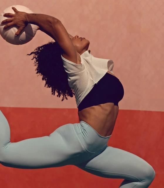 A woman in athletic wear leaps against a split pink and orange background, reaching to catch a white ball with one hand overhead.