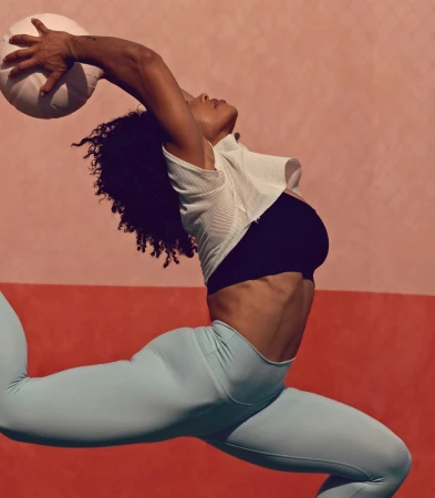 A woman in athletic wear leaps against a split pink and orange background, reaching to catch a white ball with one hand overhead.
