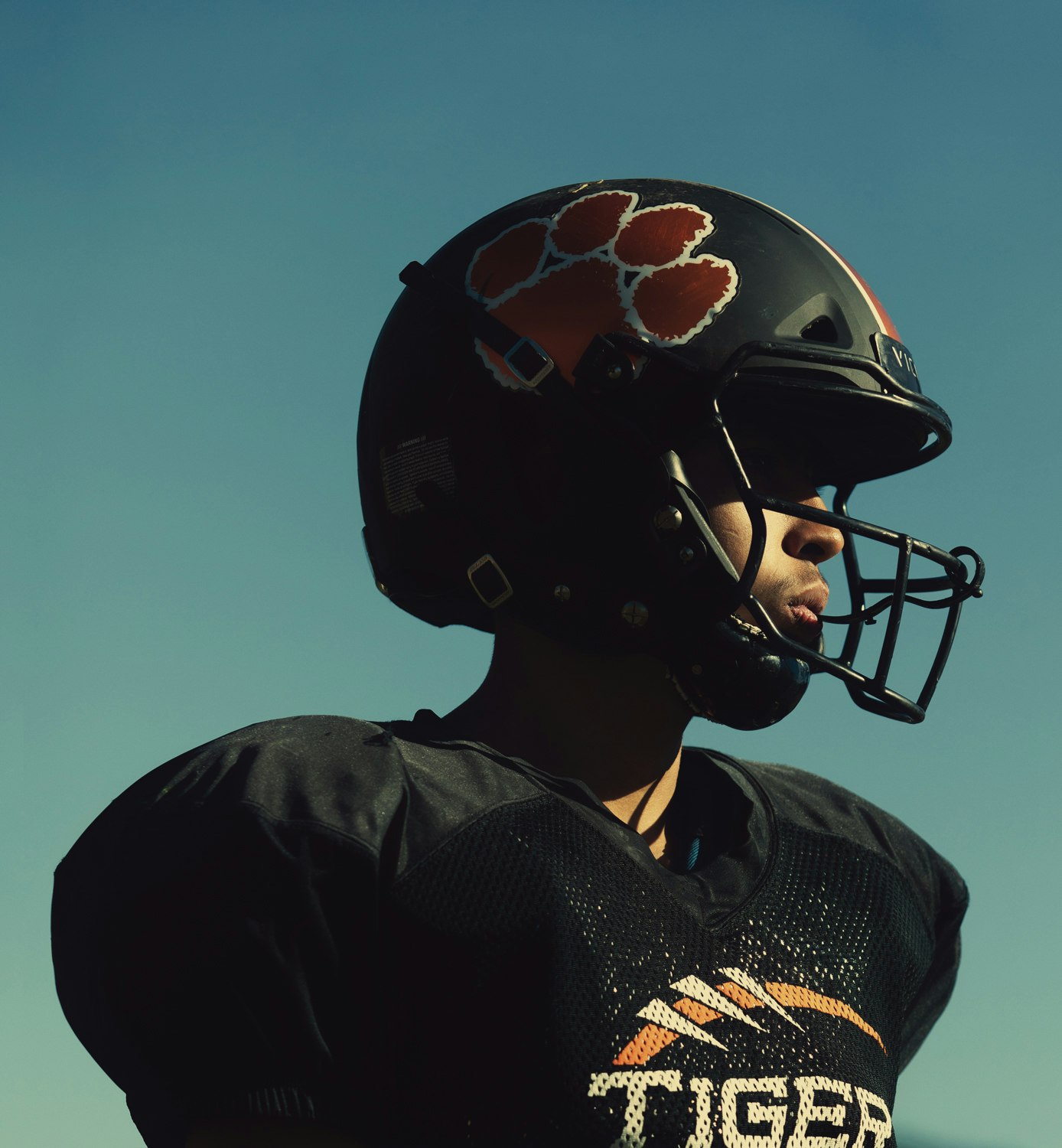 A football player in uniform and helmet looks off-camera against a clear blue sky, emphasizing the detail of the sports equipment.