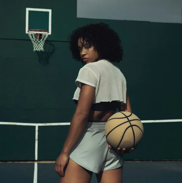 A young, curly-haired woman in athletic wear holding a basketball in an outdoor sports court environment. She looks back over her shoulder.