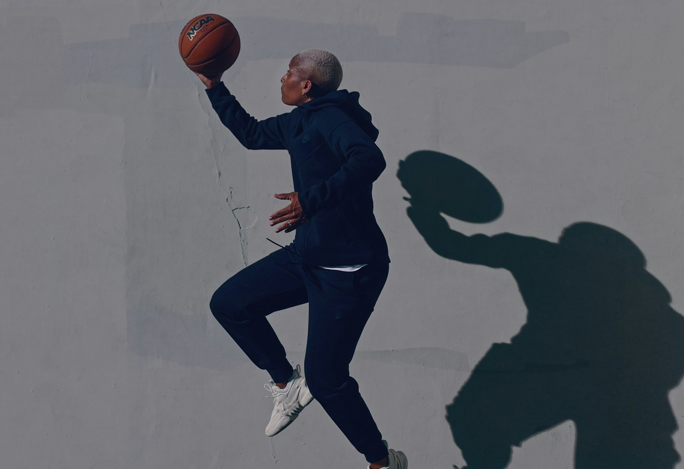 An adult female athlete leaps toward the left side of the frame with a basketball held aloft in an outdoor sports setting.