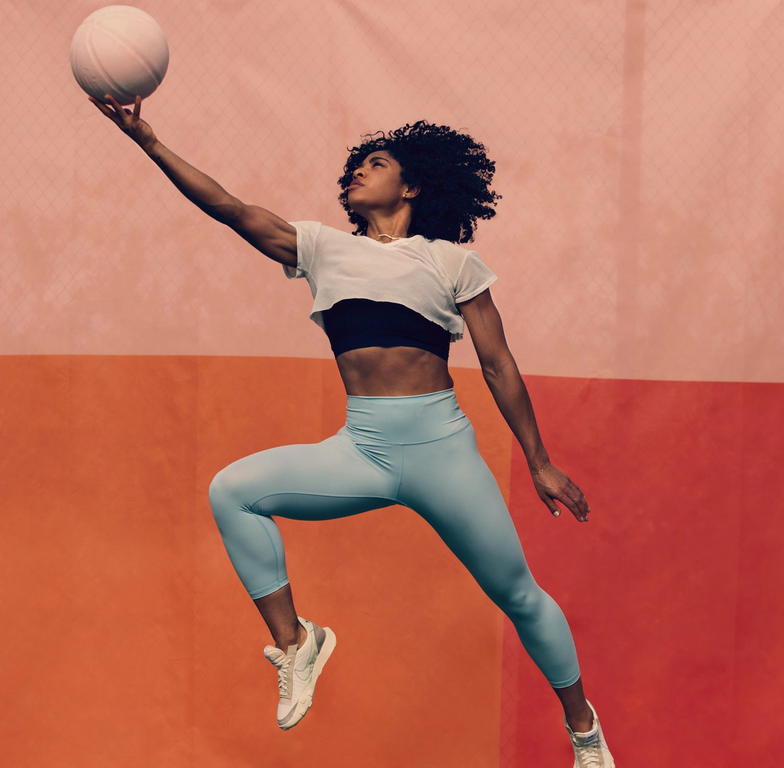 Athletic woman mid-air, jumps with a basketball, wearing athletic clothing, captured against a two-tone peach and orange backdrop.