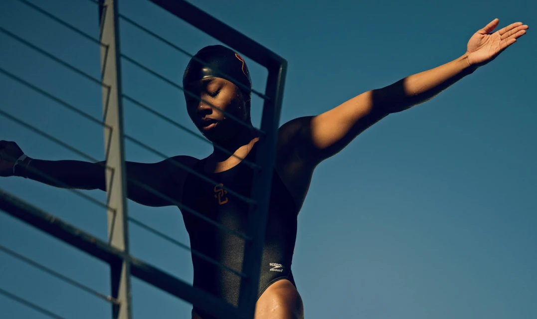 An athlete in a black swimsuit and swim cap stands with arms outstretched against a clear blue sky, framed by a metal structure.