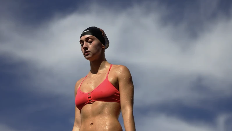 Wet female swimmer wearing a pink bikini and black swim cap against a cloudy blue sky background