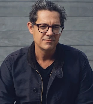 Headshot of a middle-aged man with glasses, wearing a dark jacket and black shirt, against a gray wooden wall.