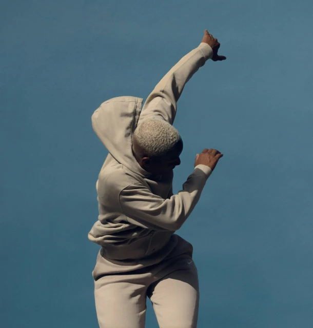 A young adult poses dynamically in beige tracksuit against a clear blue sky, with a strong emphasis on fashion and lifestyle.