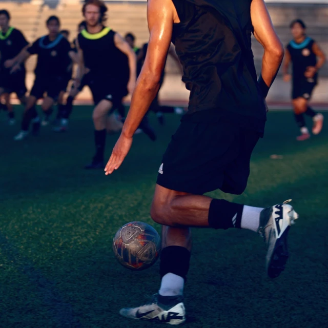 Action shot of soccer player kicking a soccer ball on a green field. Several other players and field visible in the background.