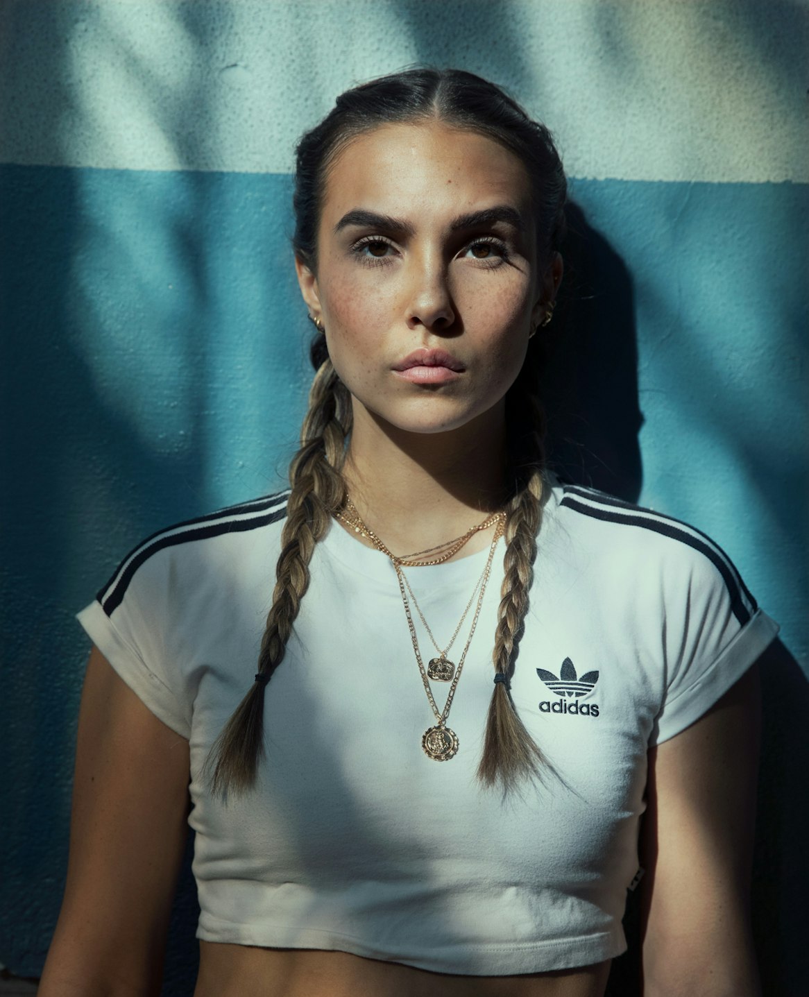 Portrait of a young woman with braided hair wearing a white Adidas crop top standing against a blue and white wall with shadow patterns.