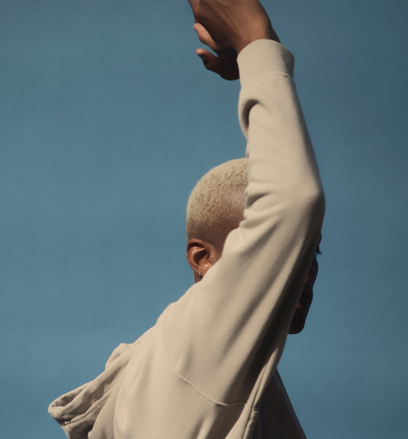Close-up of androgynous person with buzz cut, wearing beige hoodie, reaching up against light blue sky backdrop.