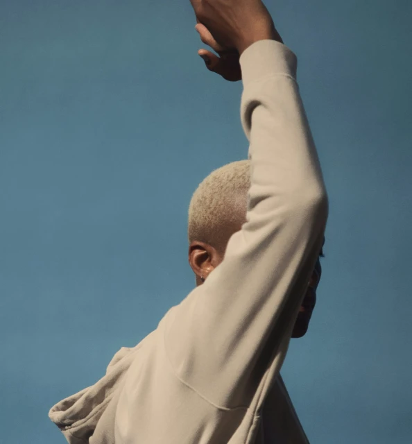 Close-up of androgynous person with buzz cut, wearing beige hoodie, reaching up against light blue sky backdrop.