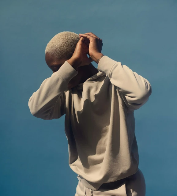 Person in a beige hoodie stands against a blue backdrop, hands near their head, showcasing a minimalistic and contemporary fashion image.