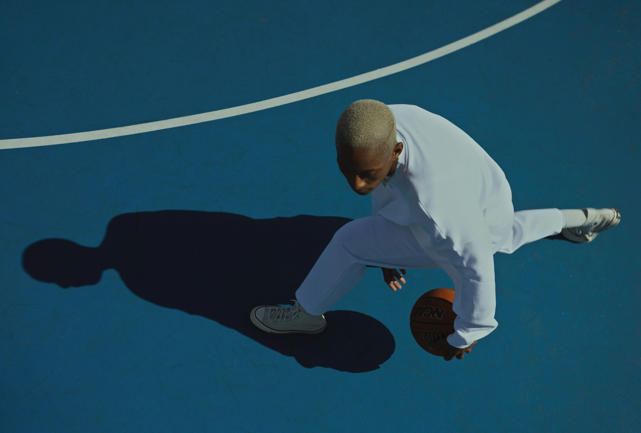 Male basketball player in white sweatsuit dribbles on blue court with white line, overhead shot casting long shadow. Athletic style.