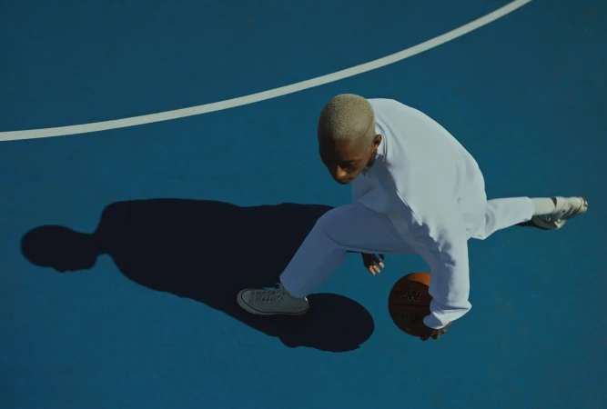 High-angle shot of a young man in white clothes dribbling a basketball on a blue court with stark shadows