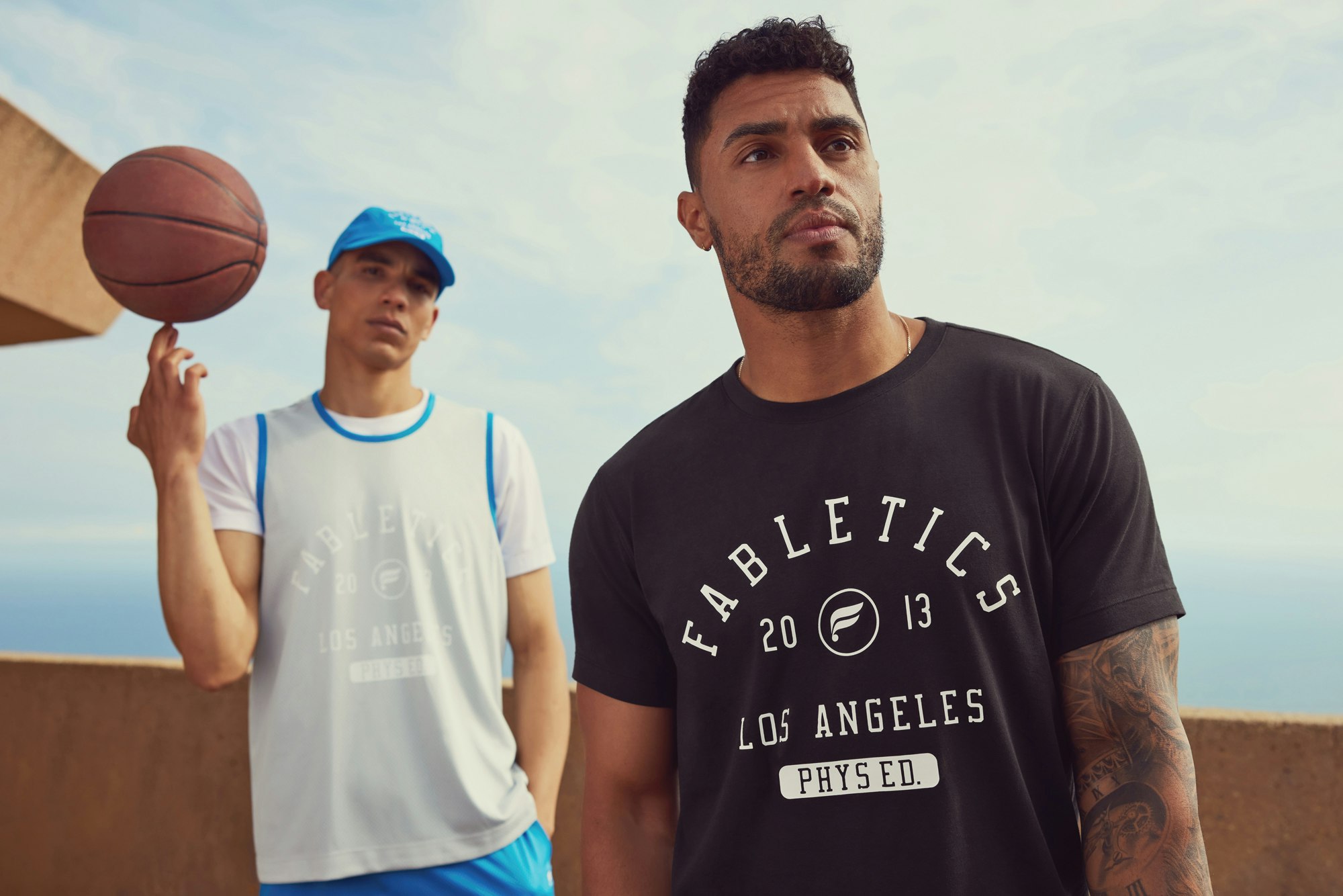 Two men in athletic wear pose outdoors with a basketball. One balances the ball on his finger, against a backdrop of sky and coastline.