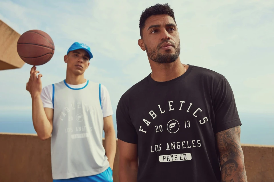 Two men in athletic wear pose outdoors with a basketball. One balances the ball on his finger, against a backdrop of sky and coastline.