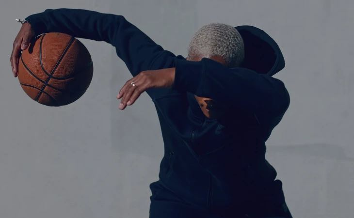 Woman with short wavy hair in a hoodie holding a basketball. Dark clothing, simple studio background, athletic attire.