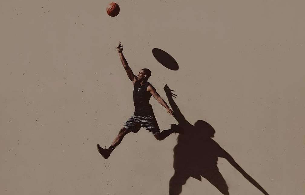 A man in a black tank top jumps to hit a basketball, casting a shadow on a tan wall in an outdoor setting, capturing action and athleticism.