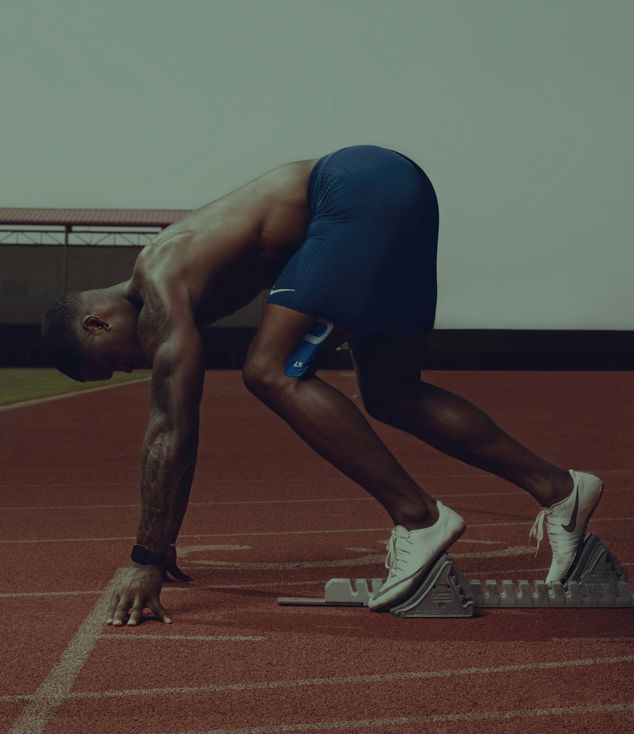 A muscular runner in navy shorts and white shoes is intensely focused in a starting block position on a red running track.