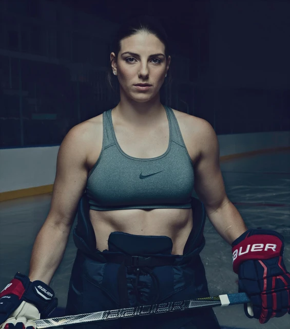Portrait of a muscular adult female hockey player on the ice rink wearing a grey sports bra and hockey gear, looking intensely at the camera.