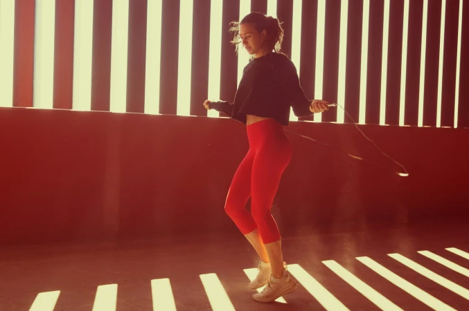 Woman wearing a black crop top and red leggings jumping rope in front of a striped illuminated wall