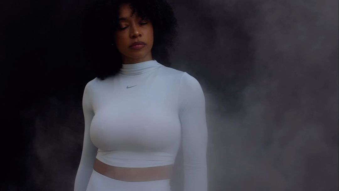 Adult female in a long-sleeve top and leggings posing in front of a dark smoky background, eyes closed, and a neutral facial expression.