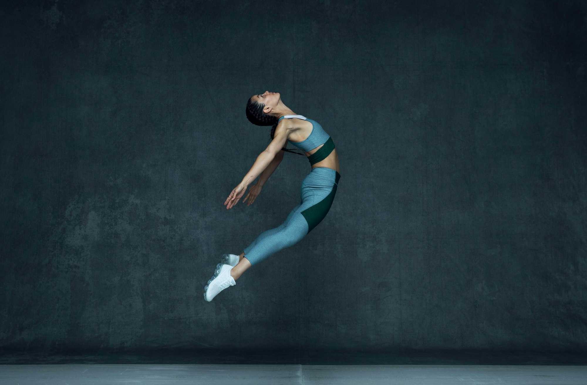 Female athlete mid-air in athletic wear against a dark textured studio wall, captured by Brian Lowe