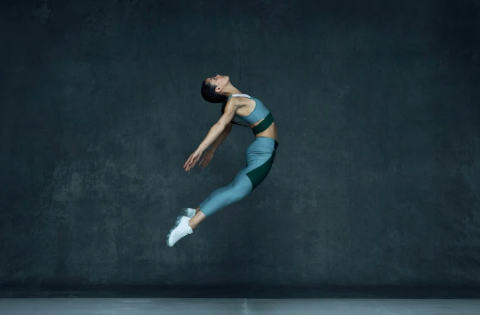 Female athlete mid-air in athletic wear against a dark textured studio wall, captured by Brian Lowe