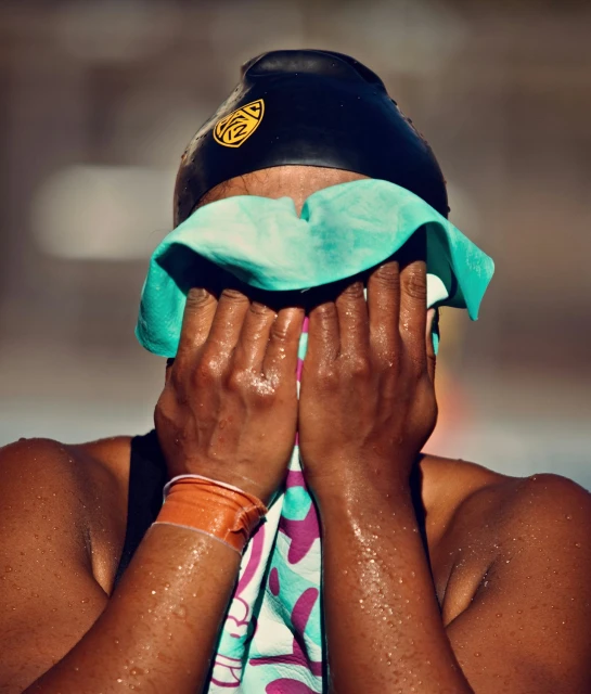 Female swimmer with wet skin in a black swim cap holding a turquoise towel to her face, shot outdoors with blurred background.