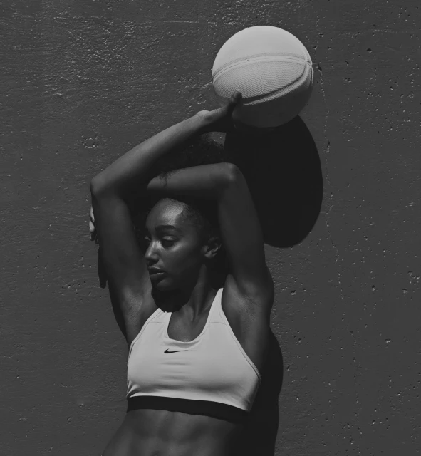 Monochrome image of a fit adult female in sportswear, spinning a basketball on her finger against a textured grey wall. Nike logo visible.