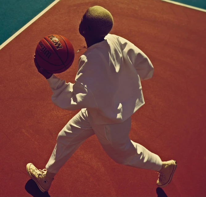 Athlete in motion on a basketball court, holding a Wilson basketball. The person wears white sweatpants, sweatshirt, and sneakers.