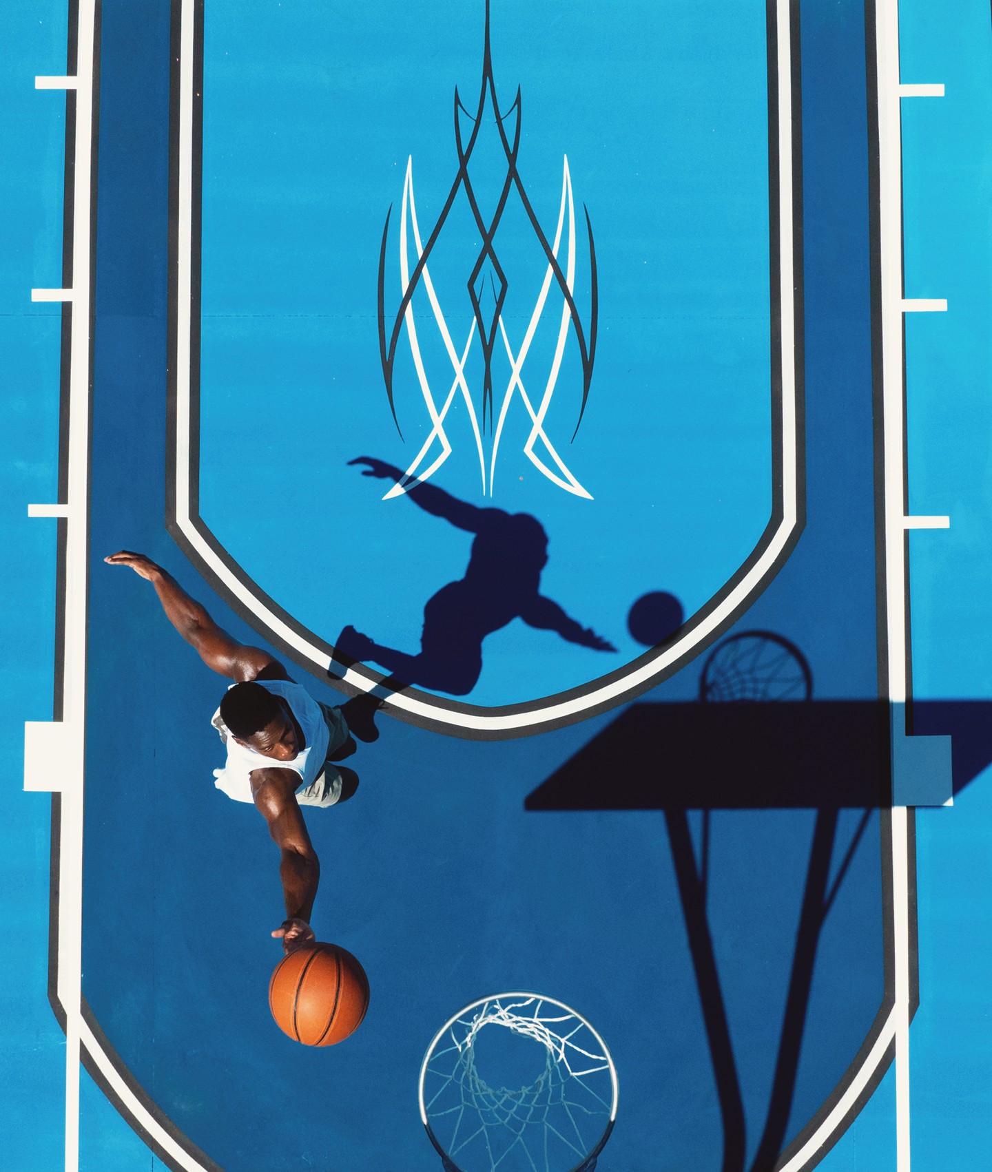 Overhead view of an adult male basketball player balancing ball on finger on blue outdoor court in full sunlight, casting a dark shadow.